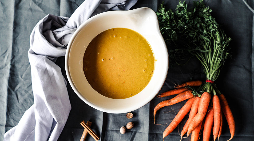 A white bowl with a pouring spout filled with a light gravy sat on a grey tablecloth with a white hand towel, a bundle of carrots, and a smattering of spices laid out next to the bowl.