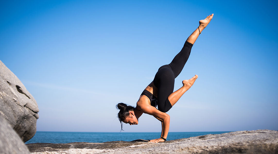 Woman doing a handstand yoga pose on a rocky foundation with blue sky and a sliver of ocean in the background.