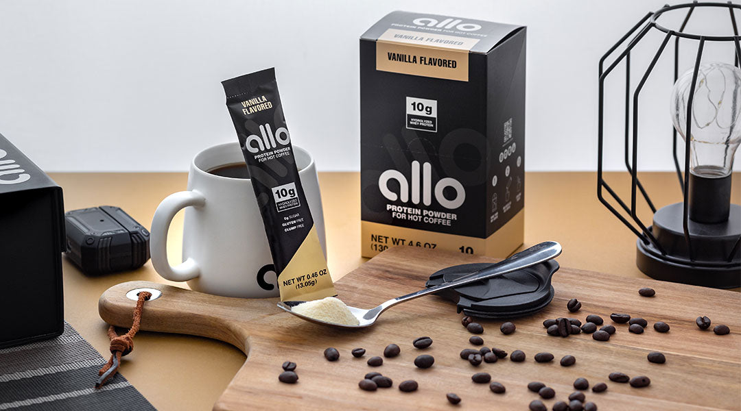 A spoon filled with protein powder and a packet and box of Allo on a table with a cup of coffee, a light, and a cutting board with coffee beans.