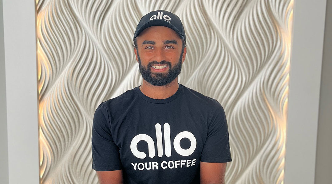 Ziggy Nathu wearing a black Allo tshirt and a black Allo baseball cap in front of a textured neutral colored wall.