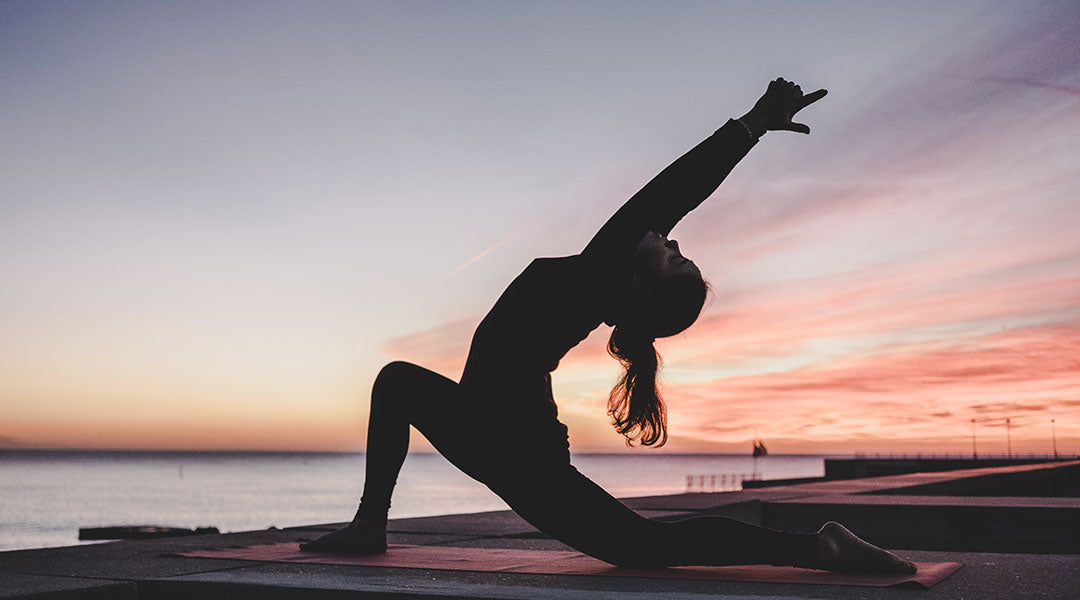 The silhouette of a woman against a waterfront sunset reaching up and back in a deep lunge yoga pose