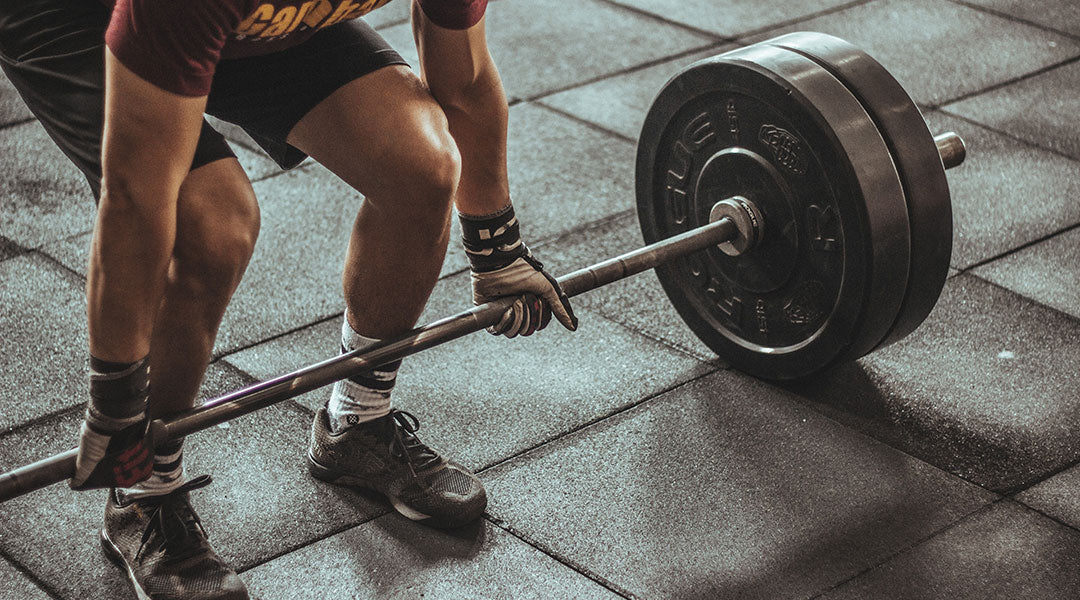 A person wearing a red shirt, black shorts, and weight lifting gloves using a mixed grip on a barbell in a deadlift start position.