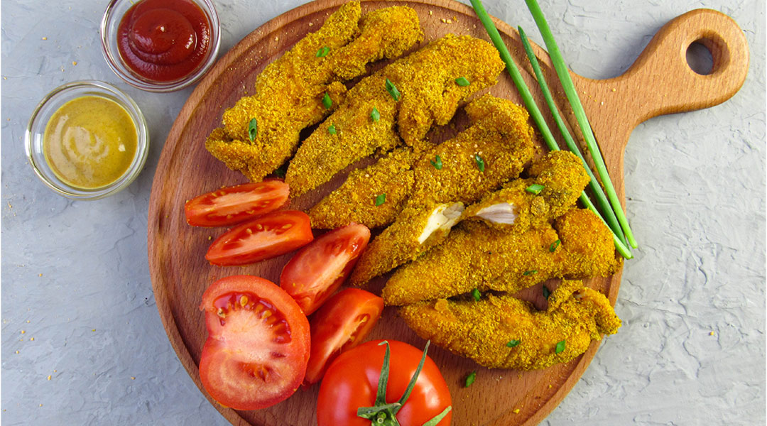 Breaded chicken tenders and sliced tomatoes on a wooden cutting board laid on a textured white and grey background with a dish each of mustard and ketchup to the side of the board.
