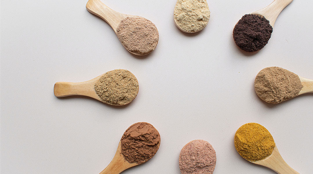 Eight identical wooden spoons laid out in a circle on a taupe background with their handles pointing out and different colors and shades of powders heaped on their heads.