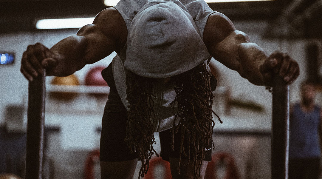 A man in a grey hoodie holds onto onto the posts of a strength training sled with his head hung so that his face is hidden but his long thing braids hand down from under the hood.