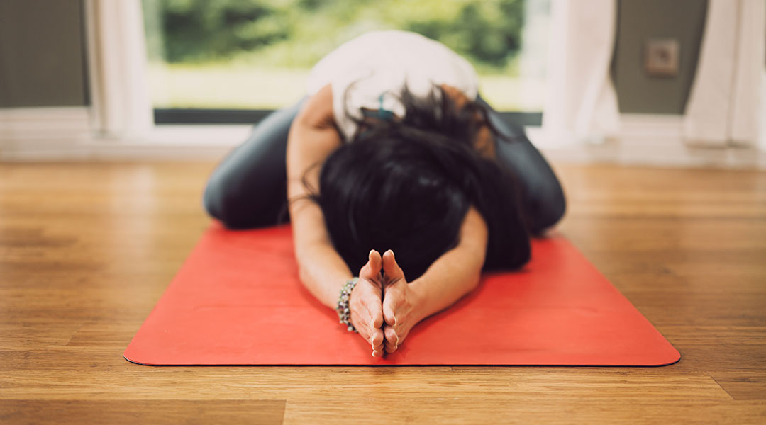 Woman in a forward bend with hands clasped overhead stretched out on an orange yoga mat on a hardwood floor.