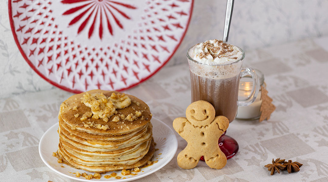 A large stack of pancakes topped with banana slices and nuts on a white plate sat next to a glass of hot cocoa and a gingerbread man on a tan table with a red and white plate in the background.