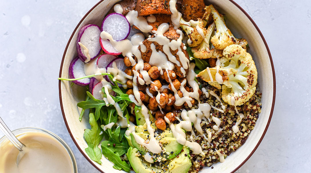 A bowl filled with colorful ingreeients like avocados, cauliflower, sweet potatoes, lettuce, and sliced radishes topped with a creamy dressing that sits to the side in a cup with a spoon all over a light grey marble background. 