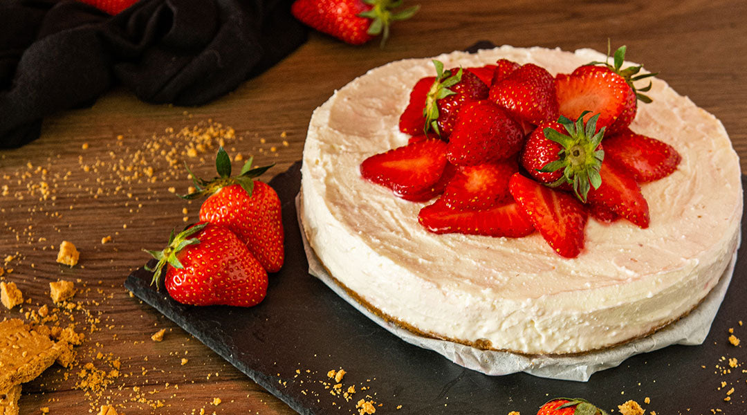 A round white cheesecake topped with strawberries on a square black plate on a wooden table covered in crumbs.