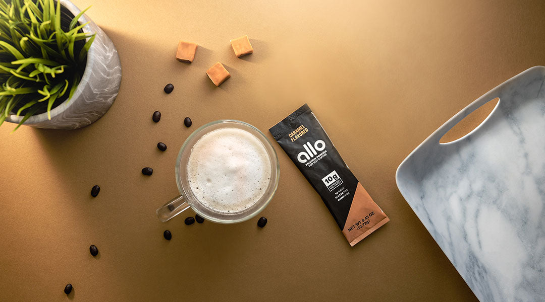 A frothy cup of coffee and packet of Allo protein powder on a dark yellow background with coffee beans, caramel cubes, a grey marble serving tray and a small potted plant. 