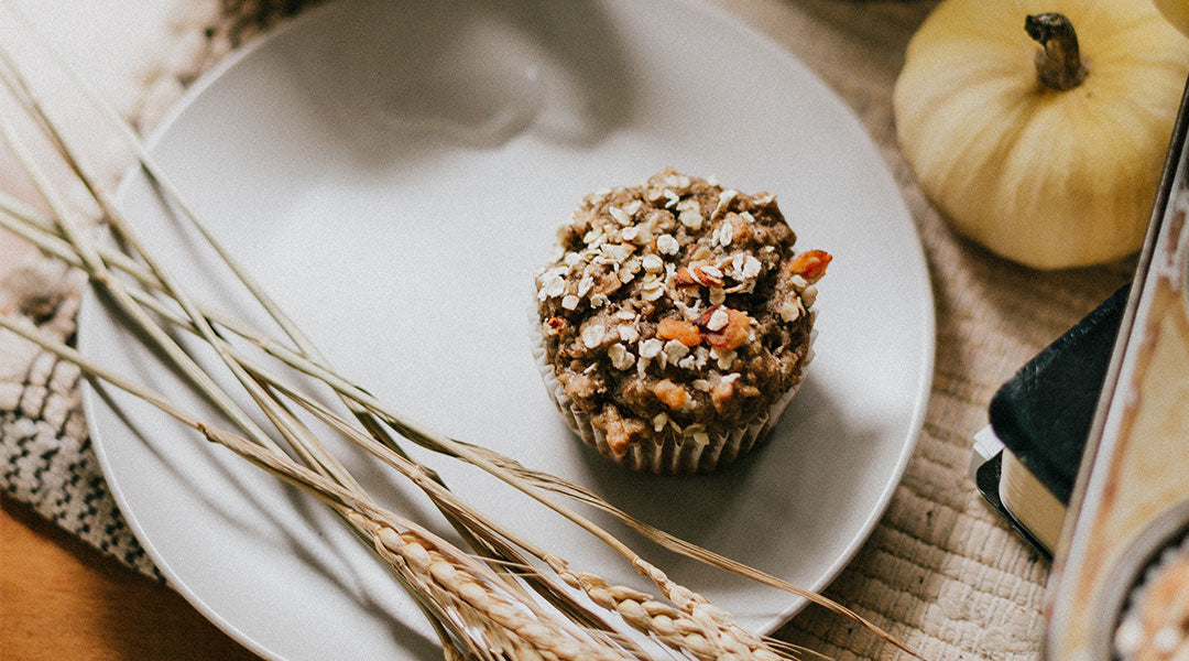 A pumpkin muffin topped with oats on a white plate with stalks of oats and a tiny yellow pumpkin of to the side on the beige placemat underneath. 