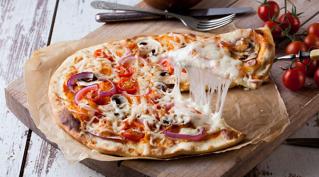 A slice of pizza being lifted away from the rest of the pie with cheese stretching between and tomatoes and cutlery in the background.