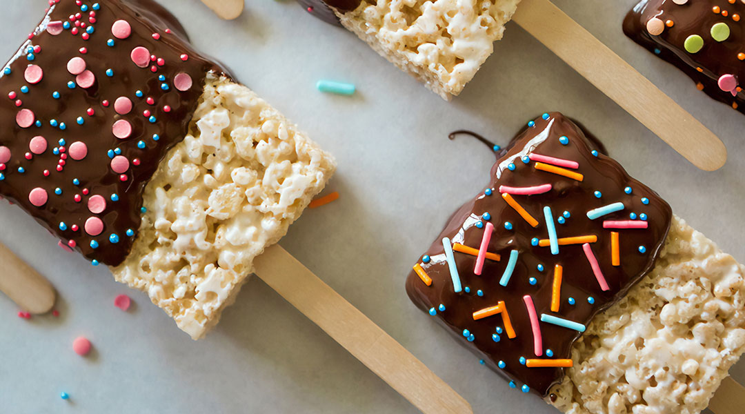 Rice crispy treats half dipped in chocolate and sprinkles on popsicle sticks 