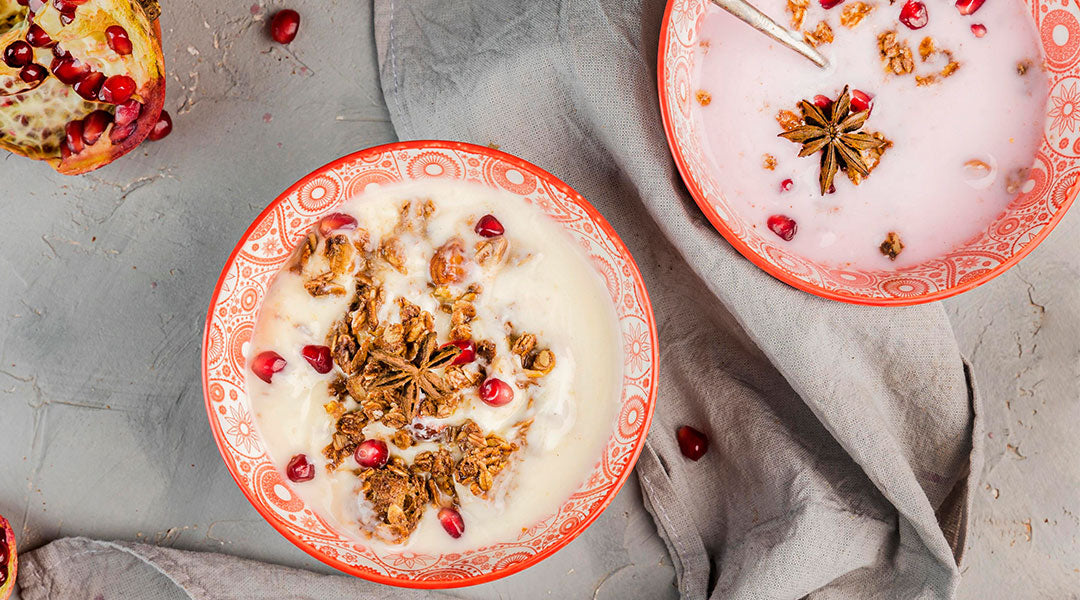Two red and white bowls filled with oats, milk, pomegranate, and star of anise set on a grey tablecloth.